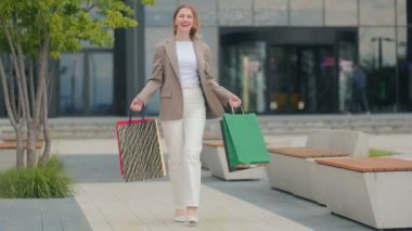 Bej ceketli, beyaz pantolonlu, kendine güveni tam bir kadın alışveriş merkezinin dışında güneşli bir günde keyifle yürüyor..