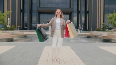 Havalı sarışın kadın, şık bir şehir binası ve çeşme meydanının önünde elleri çanta dolu, parlak bir şekilde gülümseyen güneşli bir alışveriş çılgınlığından hoşlanıyor..