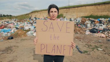 Çevre aktivisti bir kadın, bir çöp sahasının önünde durup kirlilik ve iklim değişikliğiyle ilgili eylem talep ederken gezegeni kurtarın yazan bir karton tabela tutuyor..