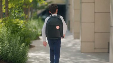 Arkadan bakan genç bir çocuk, siyah bir sırt çantasıyla okula giderken enerjik bir şekilde zıplıyor. Sahne, neşe ve hareketle dolu tasasız bir sabahı yakalar..