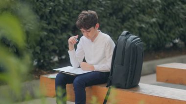 Okul bahçesindeki bankta oturan bir çocuk ev ödevini yapıyor. Defterine, sırt çantasına ve yanındaki kağıtlara yazarken ifadesi odaklanmış durumda..