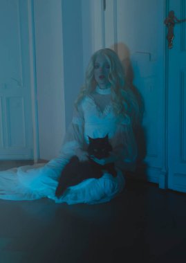 Siyah bir Maine-Coon kedisi olan beyaz elbiseli genç bir kadın yerde, bir odada oturuyor. Oda mavi ışıkla dolu. Gizemcilik.