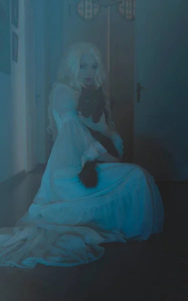 穿着老式白衣的年轻女人和一只黑猫坐在地板上的房间里 房间里充满了蓝光 神秘主义 — 图库照片