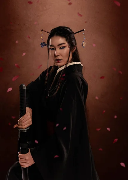 日本女武士身穿和服 手握褐色背景和花瓣的卡塔娜 日本文化 图库图片