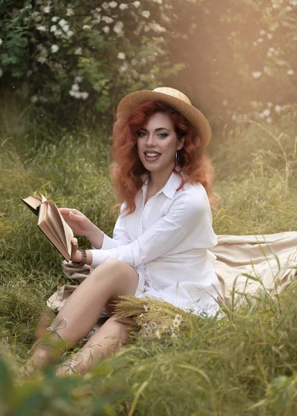 街の公園で本を読んでドレスや帽子の若い赤毛の女性 — ストック写真