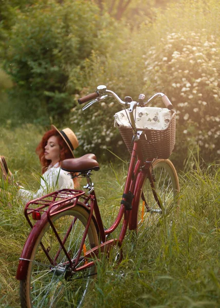 街の公園で本を読んでレトロな自転車でドレスや帽子の若い赤毛の女性 — ストック写真
