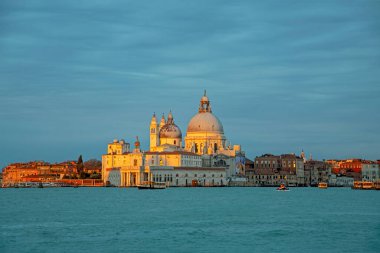 Venedik, İtalya - 27 Şubat 2023: Venedik 'te Santa Maria Della Salute Kilisesi mavi gökyüzü ve su arka planına karşı altın gün ışığı ile aydınlatıldı. Güzel karşıtlık şehir manzarası