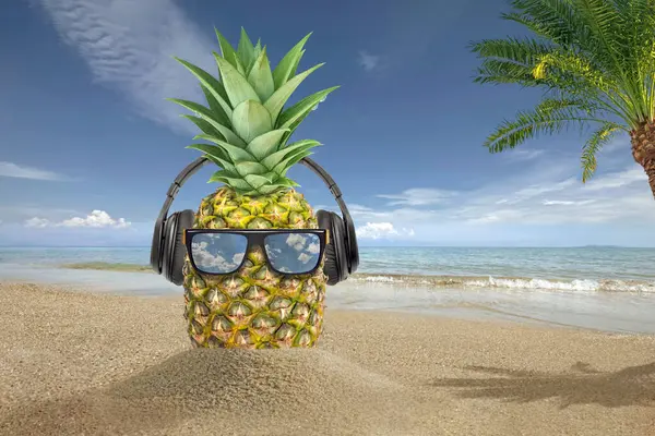 Hipster pineapple in sunglasses and listen music with headphone on the sand beach beautiful blue sky and palm tree background. Fashion, summer, vacation and party concept, copy space for banner