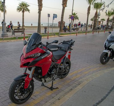 Larnaca, Kıbrıs - Nisan 01, 2024: Kırmızı Ducati Multistrada motosikleti palmiye ağaçları ve arka planda deniz bulunan bir caddeye park edildi