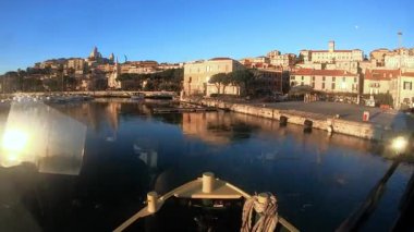 Bir balıkçı teknesi gün batımında Imperia Porto Maurizio marinasından ayrılıyor, pruvadan su ve rıhtımdaki teknelere yansıyor..