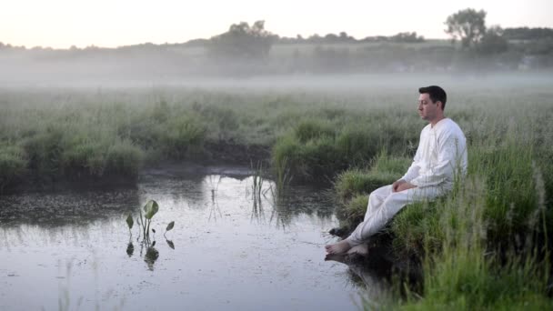 白い服を着た男が小さな森の湖のほとりの芝生の上に腰を下ろしている ビデオ — ストック動画