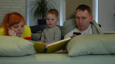 Young parents and little boy in bed. Mom and dad look at the screens of phones, the child independently reads a book aloud. Parents dont care. Childish loneliness