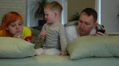 Young parents and little boy in bed. Mom and dad look at the screens of phones, the child is left to himself. The child is angry, the parents do not pay attention. child impotence