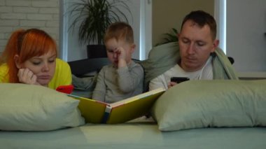 Young parents and little boy in bed. Mom and dad look at the screens of phones, the child is bored, yawns and rubs his nose. Childhood loneliness, sadness, impotence