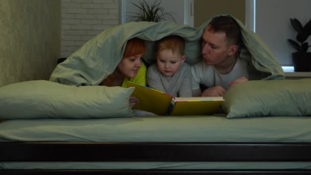 ベッドに行く前に カバーの下のベッドで若いお母さん お父さんと小さな息子 真剣な表情の子供が本を指しています ベッドで幸せな気楽な家族 全体的な計画 高品質4K映像 — ストック動画