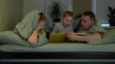 Happy young family in bed before going to sleep. Mom, dad and a small child are reading a book in bed under the covers and laughing. Daily ritual. High quality 4k footage