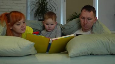 Young parents and little boy in bed. Mom and dad look at the screens of phones, the child independently reads a book aloud. Parents dont care. Childish loneliness. High quality 4k footage