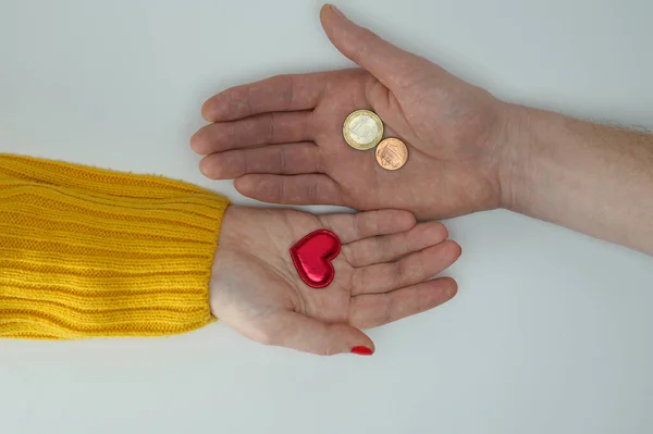 Love for money, selling love. Two hands, male and female, extend their palms to each other. In one hand is a red heart, in the other hand are coins. Close-up. Isolated on white background.