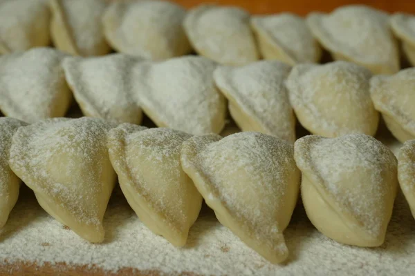 Raw handmade dumplings on a wooden kitchen board sprinkled with flour on top. Beautiful rows with dumplings. Ukrainian national dish. Close-up.