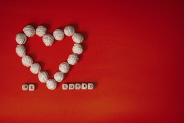 A white heart made of knitted balls and the inscription be loved from white wooden cubes on a red vintage background. Place for text.