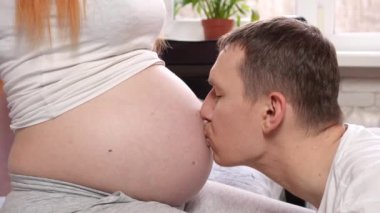 Genç müstakbel babası hamile karısının karnını öpüyor. Gelecekteki baba hamile karısının karnını okşar ve gülümser. Yakın plan. Yüksek kalite 4k görüntü
