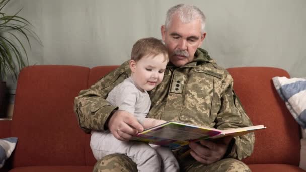 ウクライナ軍の制服を着た男が自宅のソファの上の小さな男の子に本を読みます 祖父は孫に本を読んでいる ウクライナ軍 — ストック動画