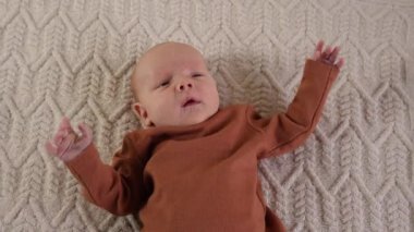 Kahverengi kazaklı küçük bir çocuk örgü örmüş bir battaniyeye uzanıyor, kollarını hareket ettiriyor ve hapşırıyor. Üst manzara, yakın plan portre. Ağır çekim. Annelik, aile, doğum, kundaklama özgürlüğü kavramı.