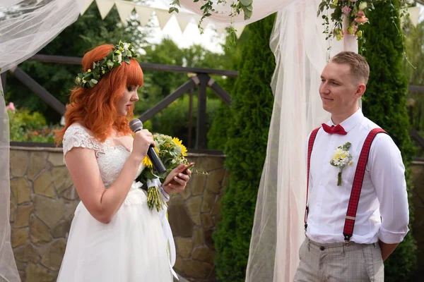red-haired bride with flowers reads from the phone into the microphone to the groom in a white shirt and red suspenders