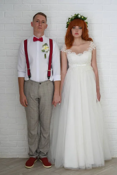 Young bride and groom with sad faces hold hands near a white brick wall. Sad newlyweds. Vertical photo.