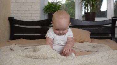 Bebek beşiğe tereddütlü bir şekilde oturur ve şakacı bir şekilde gülümser. Beyaz tulumlu küçük kulaklı sevimli bir çocuk yatağın ortasında neşeli bir ruh hali içinde. Yüksek kalite 4k görüntü