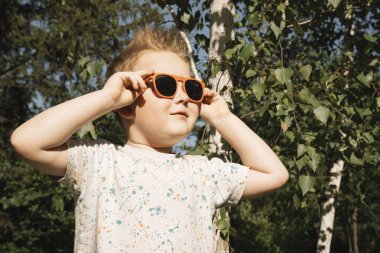 Yeşil ağaçların arkasında, güneş gözlüklü mutlu bir çocuğun portresi. Yakın plan. Gözlüklü mutlu çocuk güneşin tadını çıkarıyor. Tatil, yaz tatili konsepti. Yüksek kalite fotoğraf