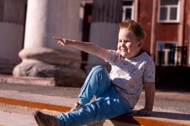 Küçük sevimli bir çocuk eski bir binanın sütunlarının arasında basamaklarda oturur ve parmağını uzaklarda bir yere doğrultur. Yüksek kalite fotoğraf