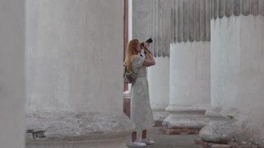 Kameralı kızıl saçlı bir kız eski sütunların arasında bir şeyin fotoğrafını çekiyor. Arkadan bak. Bir kız fotoğrafçı manzaraya bakar. Yüksek kalite 4K görüntü.
