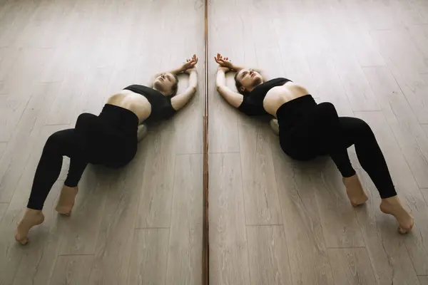 Siyah spor giyimli güzel esnek bir kız parke zeminde güzel bir şekilde yatıyor. Ayna yansıması. Sıcak bir fotoğraf. Stüdyoda jimnastik ve yoga dersleri var. Kadın vücudunun güzel kıvrımları
