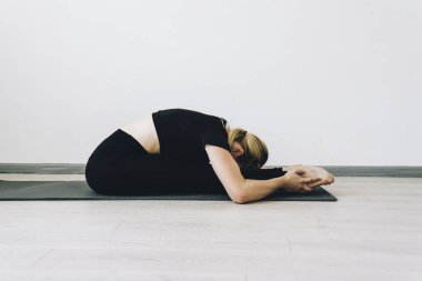Genç esnek bir kız esneme hareketleri yapar ve spor minderinde yoga yapar. Yandan ateş ediyorlar. Paschimottanasana pozu, eğitim. Stüdyoda jimnastik ve yoga dersleri var.
