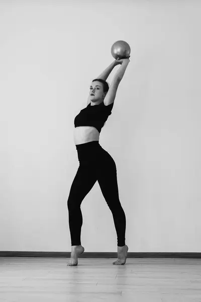 Siyah spor giyimli, beyaz bir duvara karşı elinde bir topla atletik bir kız jimnastikçi. Siyah beyaz dikey fotoğraf. Yüksek kalite fotoğraf