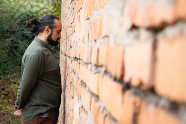 Koyu saçlı, yeşil gömlekli, uzun saçlı bir adam tuğla bir duvara bakıyor. Kolları arkasından çaprazlanmış. Yan görüş. Yüksek kalite fotoğraf