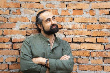 Koyu saçlı, sakallı Dominikli bir adam yeşil gömlekli, tuğla bir kırmızı duvara karşı. Duvarın yanındaki adam kollarını göğsünün üzerinde kavuşturmuş ve dikkatlice yukarı bakıyor. portre