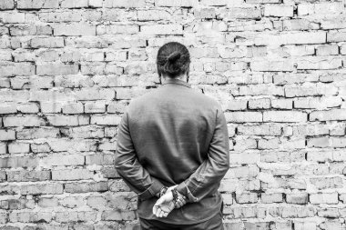 Koyu saçlı, uzun saçlı, gömlekli bir adam duvara bakıyor. Ellerini arkasından çapraz yap. Siyah beyaz, yüksek kaliteli fotoğraf.