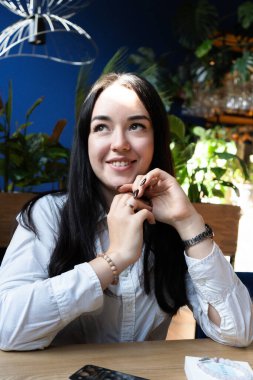 Ellerinde bilezikleri olan beyaz gömlekli güzel, koyu renk saçlı bir kız masaya oturur ve tatlı tatlı gülümser, ellerini önünde kavuşturur. Yakın plan portre. Yüksek kaliteli dikey fotoğraf