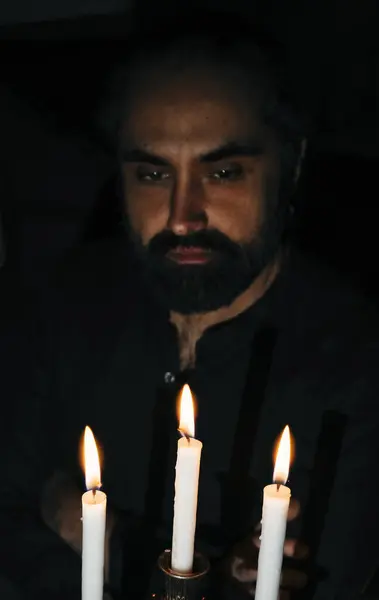 Düşünceli bir ifadeye sahip bir adam loş bir ortamda yanan mumlara bakar, sakin ve içe dönük bir atmosfer yaratır. Düşünceli tavrı derin bir yansıma olduğunu gösteriyor..
