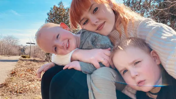 Aile açık havada güneşli bir günün tadını çıkarıyor. Bir yetişkin iki çocukla poz verir. Bir çocuk parlak bir şekilde gülümserken diğeri düşünceli görünüyor. Arka planda ağaçlar ve mavi gökyüzü. Yüksek kalite fotoğraf