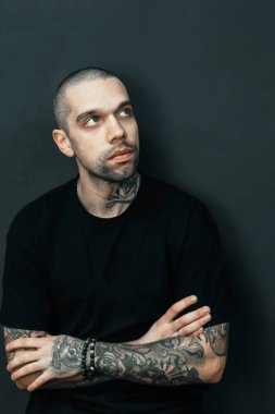 Siyah tişört giyen dövmeli adam düşünceli bir ifadeyle yukarı bakıyor. Kollarda ve boyunda görünür dövmeler, derin düşüncelere ya da karanlık bir arka plana karşı derin düşüncelere işaret ediyor. Yüksek kalite fotoğraf