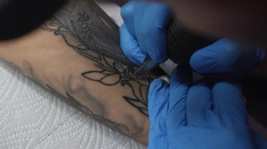 Mavi eldivenli bir dövmeci müşterinin kolunda çiçek dövmesi yaratıyor. Sanatçı tasarımın ana hatlarını çizerken dövme makinesi hareket halinde. Stüdyo ortamı