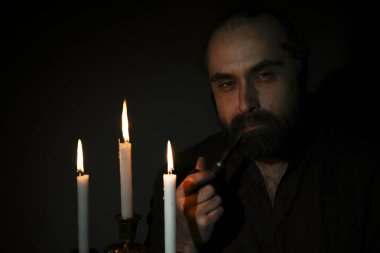 Dalgın suratlı bir adam elinde boruyla loş bir odada üç yanan mumla oturuyor. Atmosfer yansıtıcı ve sessiz, iç gözlem anını yakalıyor..