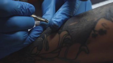 Mavi eldivenli bir dövmeci müşterinin kolunda çiçek dövmesi yaratıyor. Sanatçı tasarımın ana hatlarını çizerken dövme makinesi hareket halinde. Stüdyo ortamı