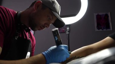 Arka planda halka ışığı olan loş bir stüdyoda müşterinin dövmesi üzerinde çalışan bir dövme sanatçısının fotoğrafını çek. Düşük açı kenar görünümü odak ve hassasiyeti gösterir yüksek kalite FullHD görüntü