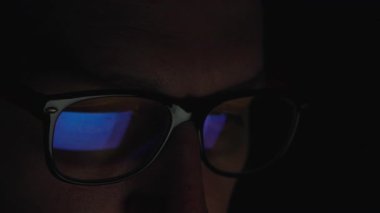 Akşamları ekranın önünde oturan bir adamın yakın çekimi. Hızlı haber yayını gözlüklerine yansıyor. Loş ışık dramatik ve yoğun bir hava yaratır. Yüksek kalite 4k görüntü