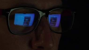 Erkek yüzü, ekrandaki kaydırmalı haber akışını yansıtan hızlı kaydırmalı haber yayını gözlüklerine yansıyor. Loş ışık dramatik ve yoğun bir hava yaratır. Yüksek kalite 4k görüntü