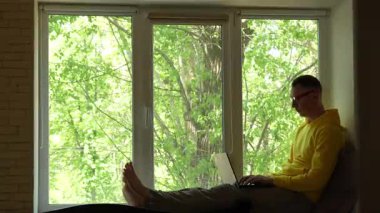 Sarı kapüşonlu ve güneş gözlüklü genç bir adam kucağında dizüstü bilgisayarla pencere pervazında oturur, çalışır ve pencereden dışarı bakar. Dışarıda yeşil ağaçlar, gün ışığı, sakin ve odaklanmış atmosfer..